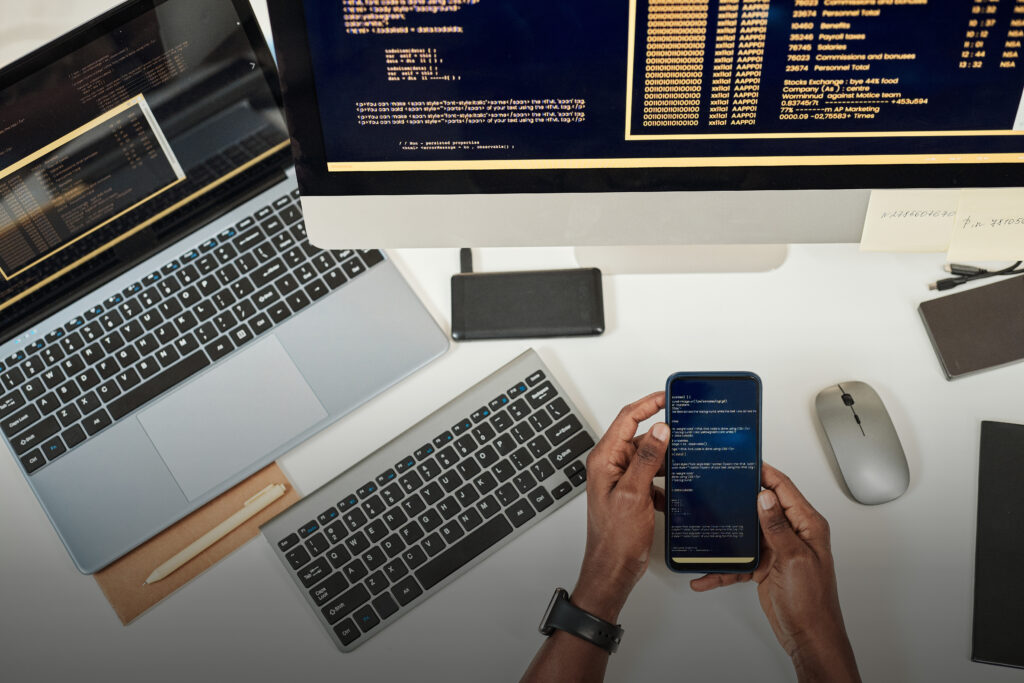 TP développeur web et web mobile - Vue du dessus, mains qui tiennent un smartphone au-dessus d'un bureau avec un ordinateur et un écran, des lignes de codes sont affichées sur tous les écrans