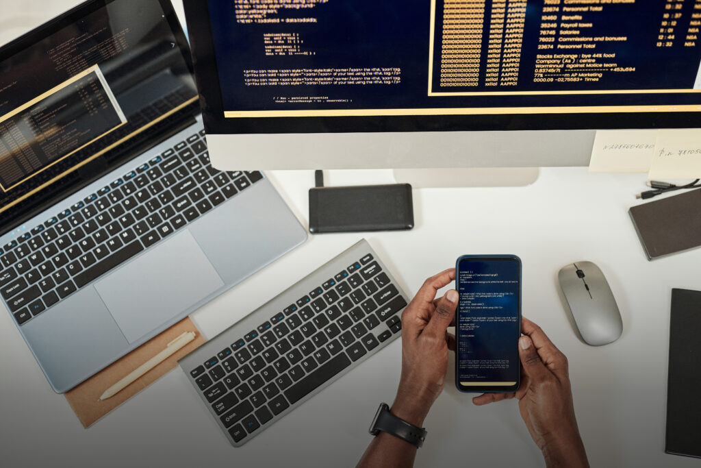 TP DWWM - Vue du dessus, mains qui tiennent un smartphone au-dessus d'un bureau avec un ordinateur et un écran, des lignes de codes sont affichées sur tous les écrans