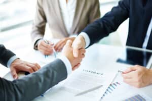 Les nouvelles techniques de recrutement -BLOG- Un gros plan sur une poignée de main entre deux personnes en costume, symbolisant la conclusion d'un accord professionnel. Un contrat signé, et des documents sont également visibles sur la table.