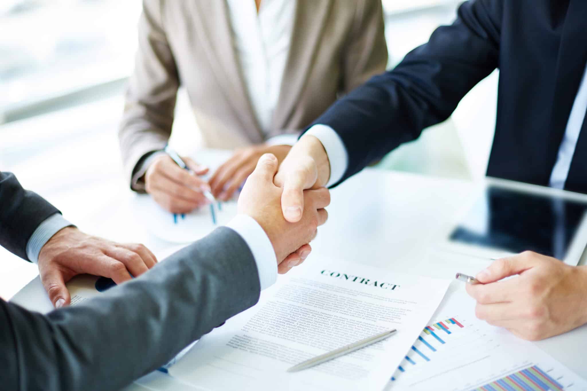 Les nouvelles techniques de recrutement -BLOG- Un gros plan sur une poignée de main entre deux personnes en costume, symbolisant la conclusion d'un accord professionnel. Un contrat signé, et des documents sont également visibles sur la table.