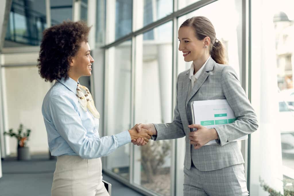 Entretenir une bonne relation avec son maître d'apprentissage -Blog- Deux femmes d'une entreprise se serrent la main dans un hall lumineux, échangeant un sourire professionnel.