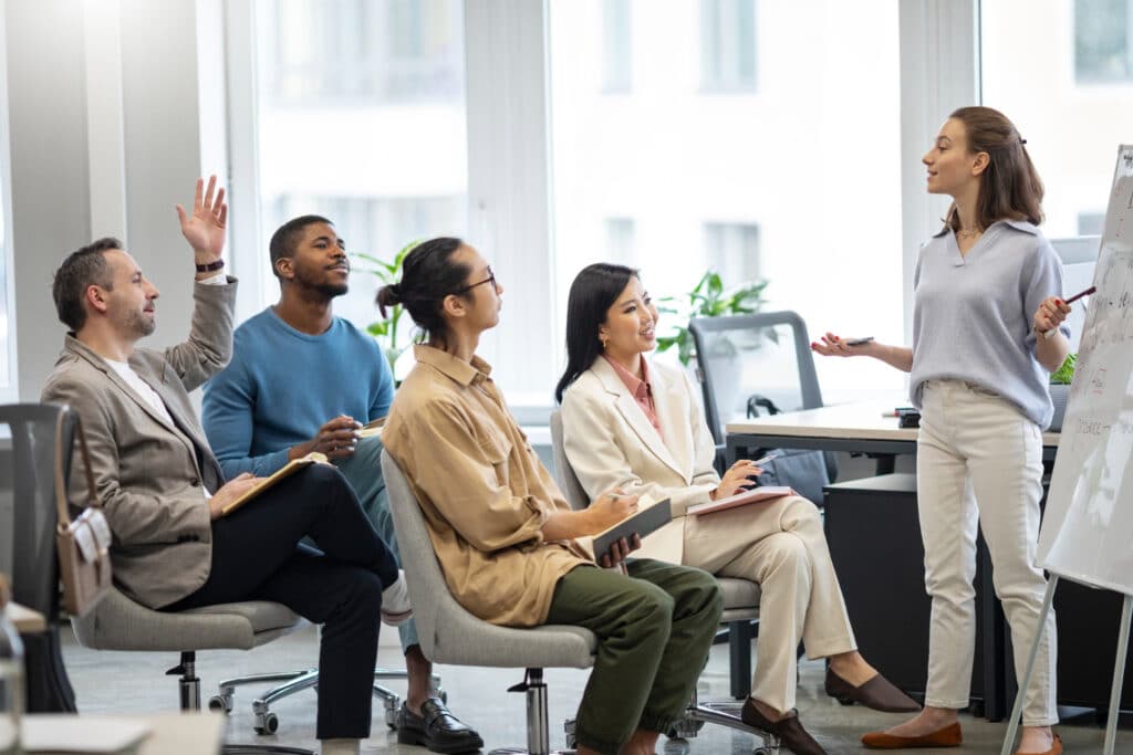 Groupe de personnes, dont quatres sont assise, et une femme debout devant un tableau blanc