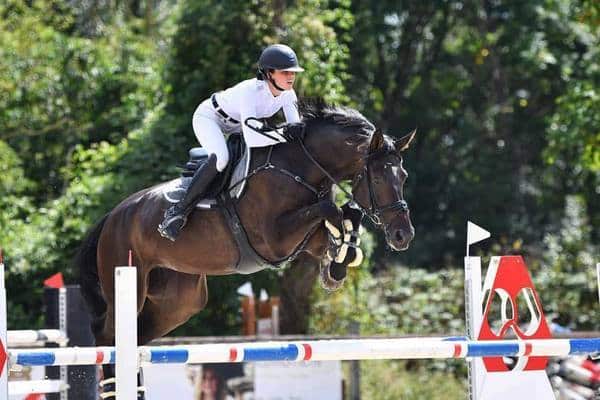 Cavalière en tenue blanche et casque noir montée sur un cheval bai foncé, en plein saut au-dessus d’un obstacle bleu et blanc lors d’une compétition de saut d’obstacles en extérieur, entourée d’arbres verts.