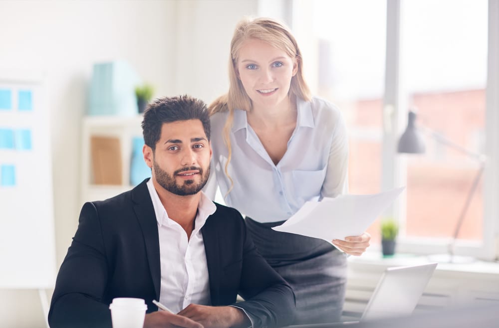 Homme en costume noir assis à un bureau avec un ordinateur portable et une femme blonde en chemise blanche debout à ses côtés, tenant des documents, tous deux souriants dans un environnement de bureau lumineux et professionnel.