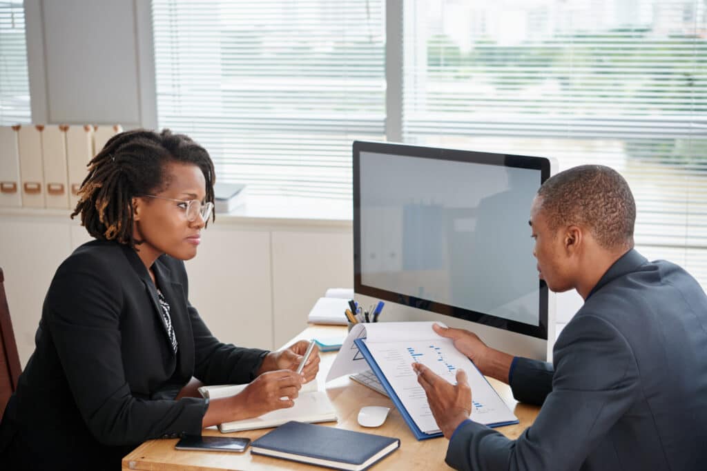 Femme à lunettes en tailleur noir assise à un bureau, discutant avec un homme en costume bleu qui lui présente un dossier avec graphiques, tous deux concentrés devant un ordinateur dans un bureau lumineux.