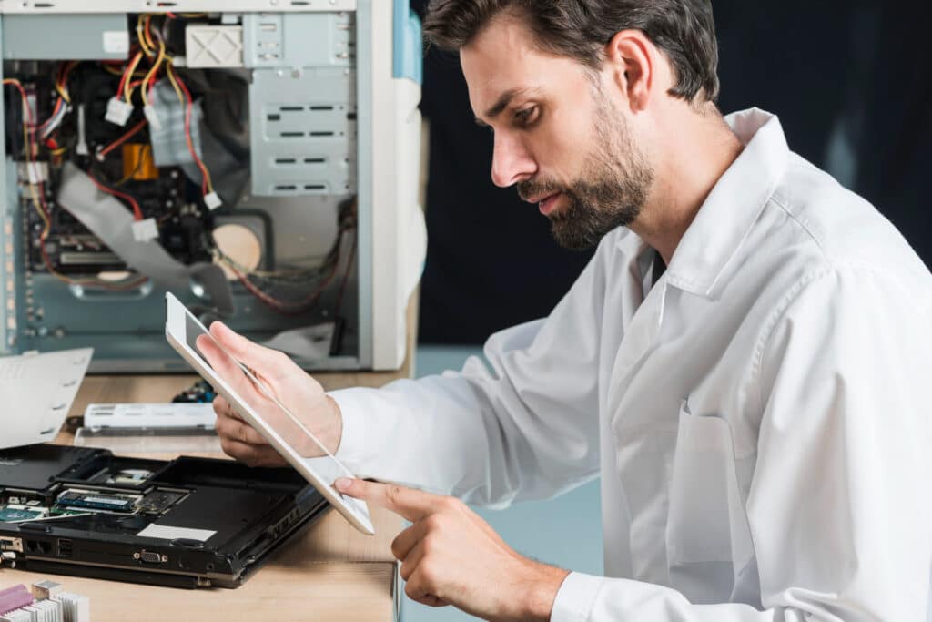 Technicien informatique barbu en chemise blanche examine attentivement une tablette tout en réparant un ordinateur portable et une tour ouverte sur son bureau, dans un atelier de maintenance high-tech.