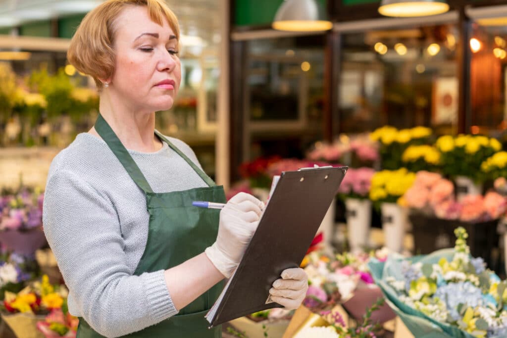 Femme fleuriste d’âge mûr en tablier vert et gants blancs, concentrée en train d’écrire sur un clipboard au milieu de sa boutique remplie de bouquets et fleurs colorées.