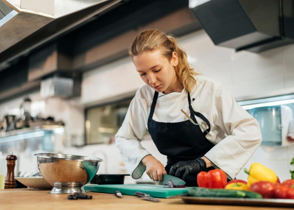 Jeune cheffe cuisinière en veste blanche et tablier noir, concentrée en train de couper des légumes sur une planche à découper dans une cuisine professionnelle équipée.