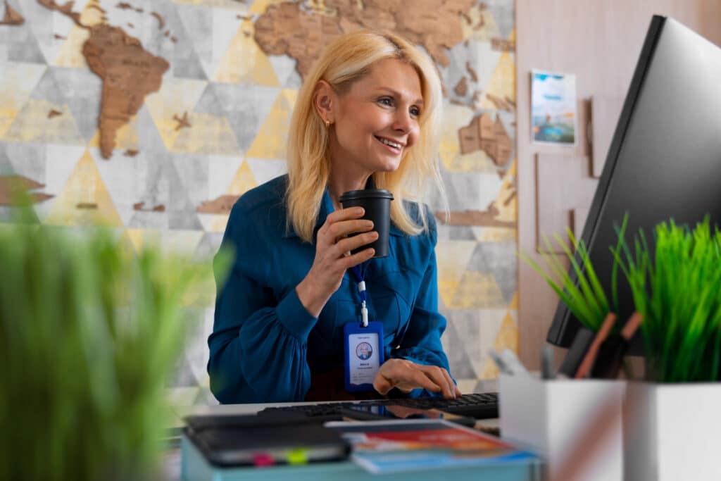 Femme blonde souriante en chemisier bleu, portant un badge, tenant un gobelet de café tout en travaillant sur son ordinateur dans un bureau moderne avec une carte du monde en arrière-plan.