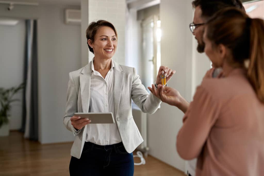 Femme en blazer gris clair souriante tenant une tablette et remettant un trousseau de clés à un couple (homme à lunettes et femme en rose) lors d’une remise de clés dans un cadre professionnel lumineux.